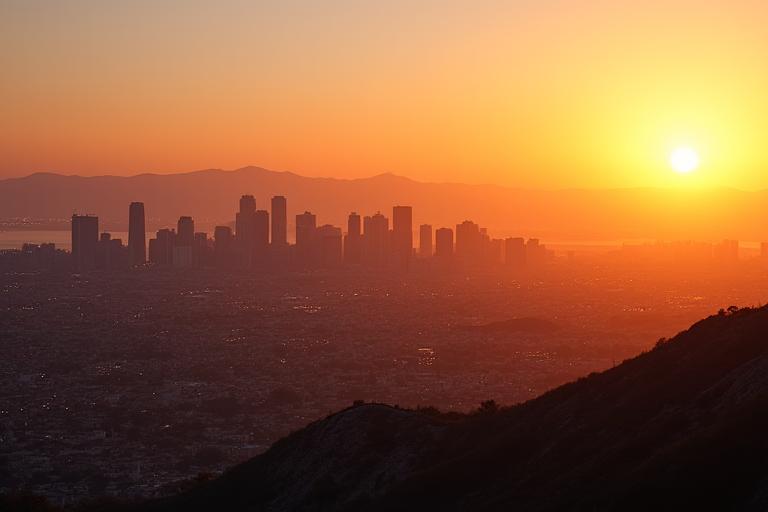Sweeping view of Los Angeles cityscape at sunset, with mountains and ocean in the distance